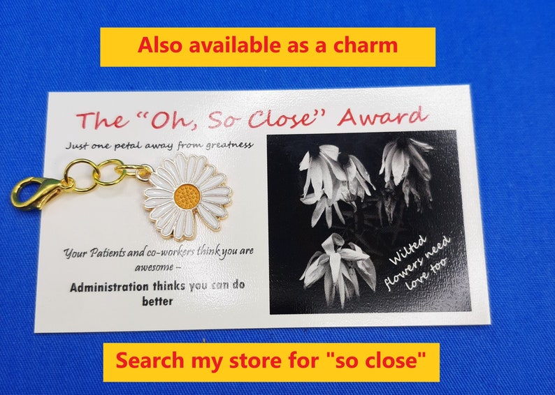 May include: A gold charm featuring a white daisy, attached to a white card with the text "The 'Oh, So Close' Award" and "Just one petal away from greatness". The card also includes a black and white photograph of wilted flowers with the text "Wilted flowers need love too".