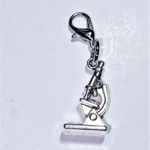 May include: Silver microscope charm with a clip on the top. The charm is a detailed miniature replica of a microscope.