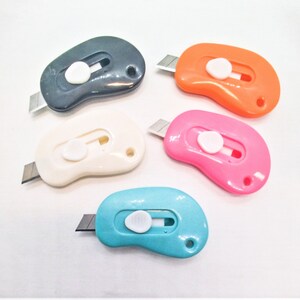 May include: Five colorful retractable utility knives with a rounded, bean-shaped design. The knives are black, white, orange, pink, and blue.