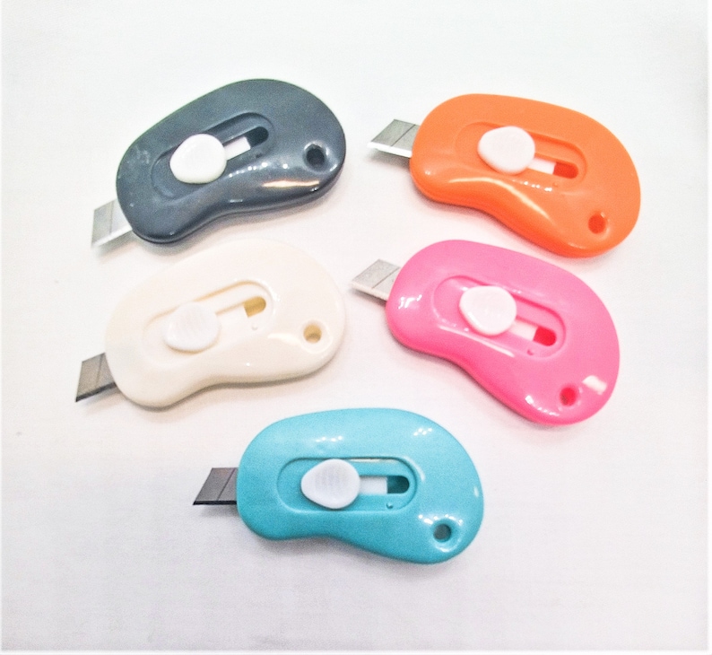 May include: Five colorful retractable utility knives with a rounded, bean-shaped design. The knives are black, white, orange, pink, and blue.