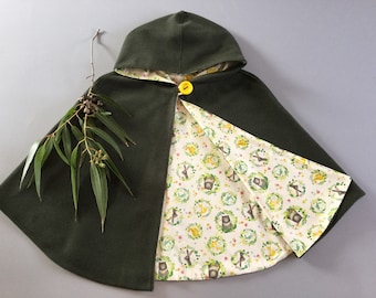 May include: A green hooded cape with a yellow button. The inside of the cape is lined with a white fabric with a repeating pattern of woodland animals and wreaths.