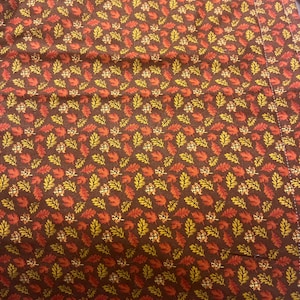 May include: Brown fabric with a repeating pattern of autumn leaves in shades of orange, yellow, and brown. The leaves are densely packed, creating a textured appearance. The fabric appears to be a tablecloth or similar textile.