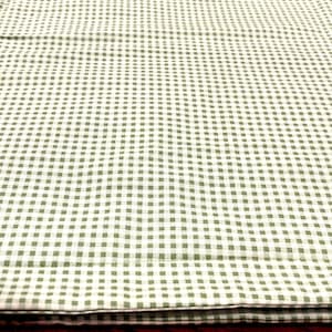 May include: A folded piece of light green and white gingham fabric. The pattern features small, evenly spaced squares. The fabric rests on a dark wooden surface. It is likely a tablecloth or similar textile.