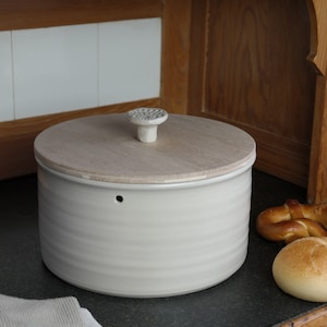 Könnte beinhalten: Eine weiße Brotdose aus Keramik mit einem Holzdeckel und einem dekorativen Knopf. Die Brotdose hat eine runde Form und eine strukturierte Oberfläche.