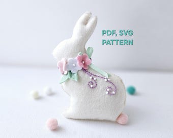 Può includere: Un coniglietto di feltro bianco con un colletto floreale e una coda a pompon rosa. Il coniglietto è decorato con fiori di feltro rosa, azzurri e verdi e un viticcio ricamato viola. Il testo sull'immagine recita "PDF, SVG PATTERN".