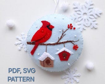 May include: A felt ornament featuring a red cardinal perched on a branch with red berries. Two small birdhouses hang from the branch. The ornament is on a white background with snowflakes.