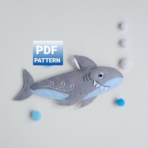 May include: A gray felt shark with light blue accents, white teeth, and embroidered details. The shark has a white ribbon and is surrounded by white and blue pom-poms. The image includes the text "PDF PATTERN".