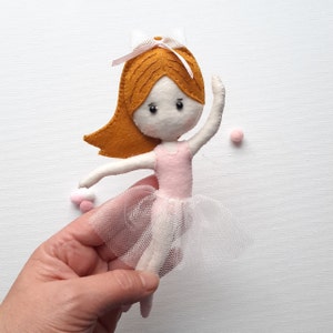 May include: A felt ballerina doll with brown hair, a pink leotard, and a white tulle skirt. The doll is holding her arm up in the air and is wearing a white headband with a pink bow.