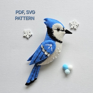 Peut inclure: Patron PDF et SVG pour un oiseau bleu avec des accents blancs. L'oiseau est fait de feutre et a un bec et des yeux noirs. Le patron comprend des flocons de neige et des pompons.