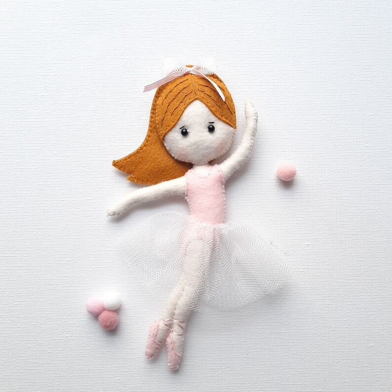 May include: A felt ballerina doll with brown hair, a pink leotard, and a white tulle skirt. The doll is wearing pink ballet shoes and has a pink bow in her hair.