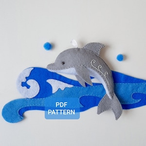 Peut inclure: Un dauphin en feutre gris sautant par-dessus une vague en feutre bleu. La vague a une crête en feutre blanc. Le texte "PDF PATTERN" est imprimé en blanc sur un rectangle rouge.