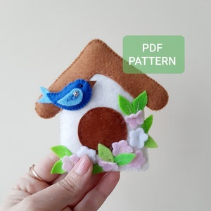 May include: A felt birdhouse ornament with a blue bird, brown roof, white walls, and pink and white flowers. The text "PDF PATTERN" is on a green rectangle.