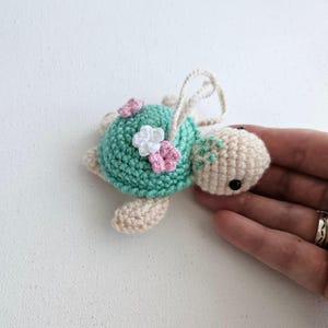 May include: A crocheted turtle ornament with a light green shell adorned with pink and white flowers. The turtle's head and flippers are beige, and it has black eyes. A loop of string is attached for hanging.