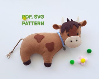 Peut inclure: Une vache en feutre faite à la main, de couleur marron et beige, ornée d'un collier en ruban bleu et d'une petite cloche. La vache a des taches marron et une frange de cheveux brun foncé. Le texte "PDF, SVG PATTERN" est en vert. Des pompons décoratifs verts et roses sont également présents.