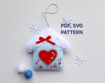 Puede incluir: Un adorno de fieltro en forma de casa azul claro con techo blanco, un corazón rojo aplicado y un lazo rojo. El adorno tiene cuentas blancas y rojas, un cordón plateado y el texto PDF, SVG PATTERN. Copos de nieve blancos y dos pequeñas bolas de fieltro están cerca.
