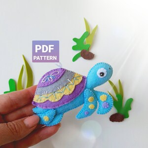 Op de afbeelding: Een vilten schildpad speelgoed met een blauw lichaam, een paarse schild en gele accenten. De schildpad heeft een grijs schild met een paars en geel bloemenontwerp. Het speelgoed houdt een PDF-patroon vast.