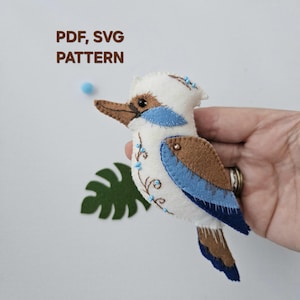 May include: A felt Kookaburra bird with white, brown, and blue accents. The bird has a detailed design with blue floral embroidery and is labeled "PDF, SVG PATTERN". A green leaf is in the background.
