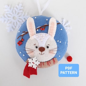 Puede incluir: Un adorno navideño de fieltro con un conejito blanco con una bufanda roja y un copo de nieve. El conejito tiene un fondo azul con cuentas blancas y bayas rojas. El adorno está etiquetado como "PDF PATTERN".