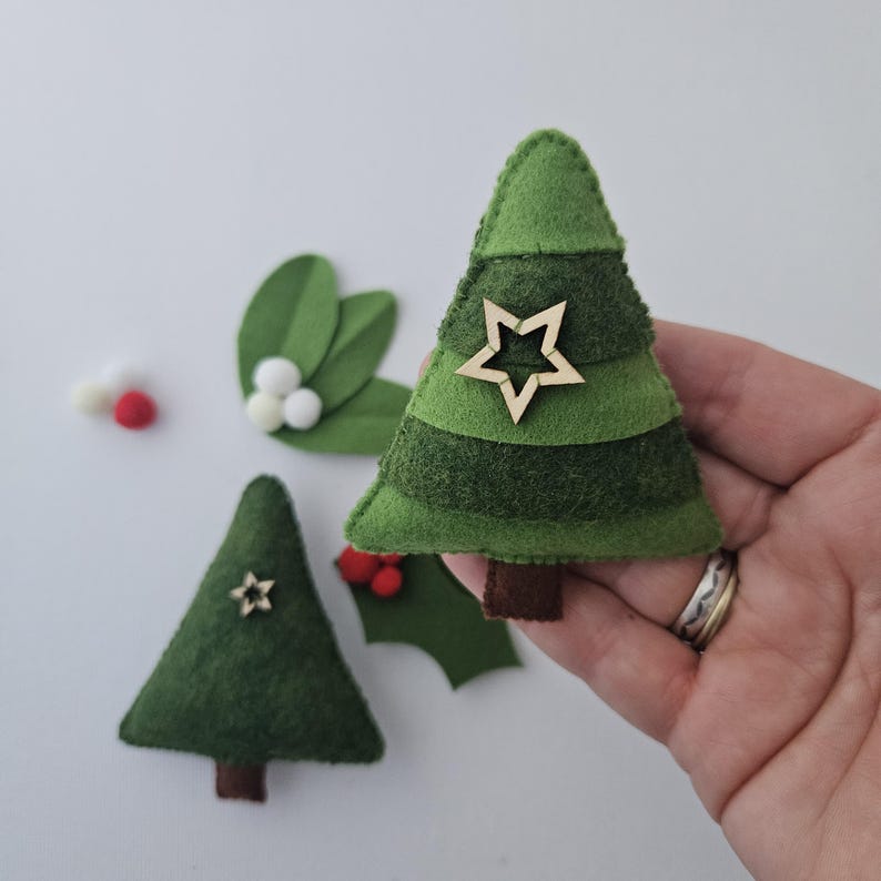 May include: Handmade felt Christmas tree ornaments in various shades of green, with a wooden star embellishment. The image also shows felt holly leaves, red berries, and white pom-poms, all on a white background. These festive decorations are perfect for the holiday season.