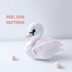 Puede incluir: Un cisne de fieltro blanco con alas de fieltro rosa y una perla en el pecho. El cisne está bordado con un diseño floral. El texto "PDF, SVG PATTERN" está escrito sobre el cisne.