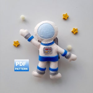 May include: A felt astronaut toy with a blue and white spacesuit, a blue helmet with a white visor, and a small red rectangle on the chest. The astronaut is surrounded by yellow felt stars and white pom-poms. The text "PDF PATTERN" is on a blue rectangle.