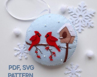 May include: A felt ornament featuring two red cardinals perched on a branch with red berries. A small birdhouse with a white roof sits on a white post in the background. The ornament is on a light blue background with white snowflakes and beads.