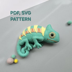 以下が含まれることがあります： 黄緑色のフェルト製カメレオンのおもちゃ。黄色とオレンジ色のアクセントと詳細な目があります。「PDF、SVG PATTERN」の文字が左上にあります。緑、ピンク、黄色の小さなフェルトボールがいくつか散らばっています。