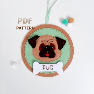 May include: A felt ornament featuring a brown and black pug with a white bone and the word "PUG" embroidered on it. The ornament is green and brown and has a loop for hanging.