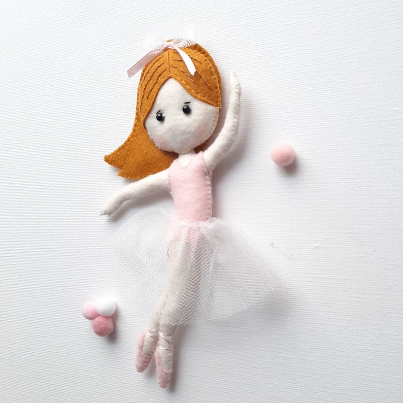 May include: A felt doll dressed as a ballerina with a pink bodice and white tulle skirt. The doll has brown hair, a pink bow, and black eyes. The doll is posed with one leg extended and the other bent at the knee.