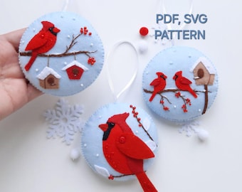 May include: Three felt ornaments featuring red cardinals on a blue background with white snowflakes. The ornaments are round and have a white string for hanging. The text "PDF, SVG PATTERN" is visible in the image.