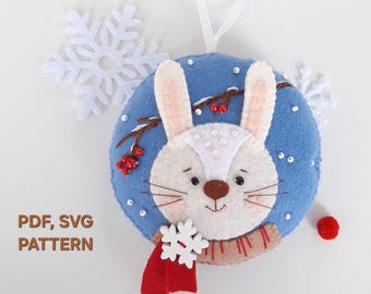 May include: A round felt ornament featuring a white rabbit face with a brown nose, whiskers, and a red scarf. The background is blue with white snowflake cutouts, red berries, and white beads. Text reads: PDF, SVG PATTERN.