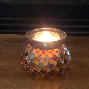 May include: A lit votive candle in a mosaic glass holder. The holder is a rounded shape with a decorative metal rim. The mosaic is made of small, colorful, square tiles in shades of gold, orange, and blue. The candle flame glows brightly.