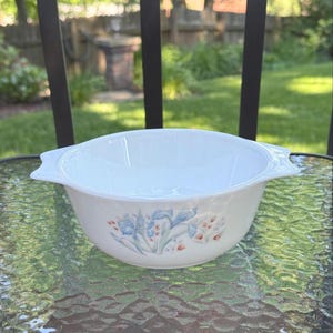 May include: A white glass bowl with two side handles, featuring a delicate floral pattern on the exterior. The design displays light blue iris flowers and small orange buds arranged in a cluster against the smooth, opaque white surface of the bowl.