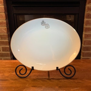May include: White oval serving platter with a gold rim and a silver crest design. The platter is on a black metal stand.