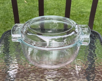 May include: A clear glass casserole dish with a matching lid and integrated side handles. The dish features a circular design with a smooth, transparent finish. The text Pyrex and Made in France are embossed on the side handles of the vessel.