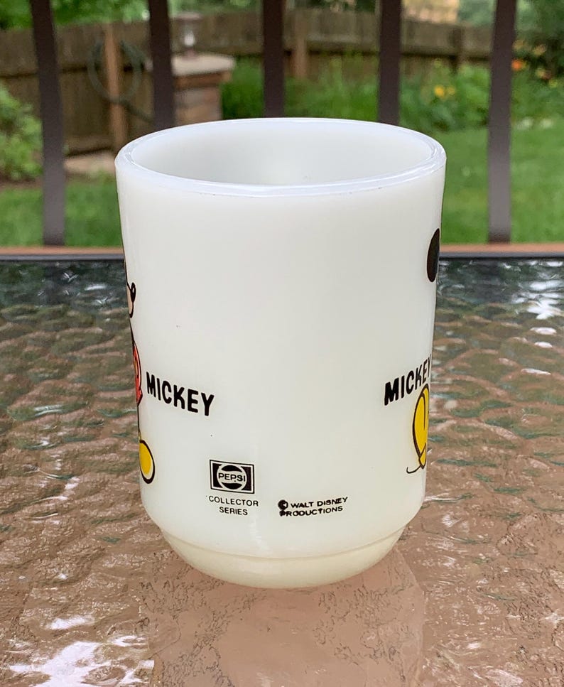 May include: White ceramic mug with a black and red Mickey Mouse illustration on one side and a black and yellow Mickey Mouse illustration on the other side. The mug has the text "Mickey" on both sides. The mug also has a black and white Pepsi Collector Series logo and a black and white Walt Disney Productions logo.