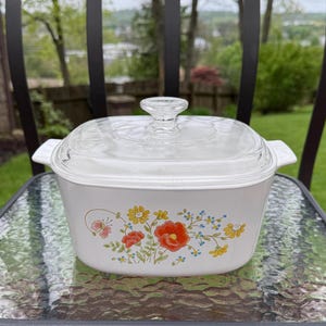 May include: White casserole dish with a clear glass lid. The dish features a floral design with orange, yellow, and blue flowers. The dish is sitting on a glass table outside.