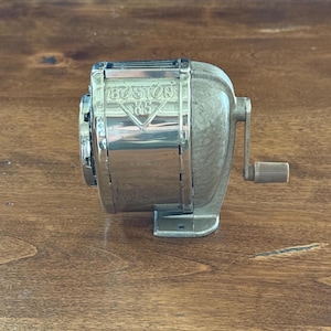 May include: Vintage Boston KS pencil sharpener. The sharpener is made of metal with a silver finish and a crank handle. The name "BOSTON KS" is visible on the front. The sharpener is sitting on a wooden surface.