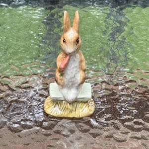 May include: A ceramic figurine of a brown and white rabbit holding a carrot. The rabbit is standing on a small, light green bench, which is placed on a bed of yellow grass. The figurine has detailed facial features and is a decorative item.