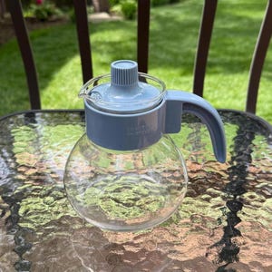 May include: A clear glass water pitcher with a round body and a light blue lid and handle. The lid has a small spout and a ribbed top. The handle is curved. The text "DO NOT USE ON STOVE" is visible on the pitcher.