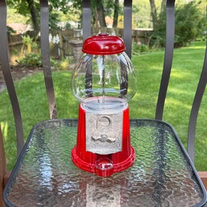 May include: A red gumball machine featuring a clear glass globe, a shiny red lid, and a silver-toned metal coin mechanism plate with an ornate, embossed floral pattern. The base is circular and red, matching the lid, with a small dispensing chute.