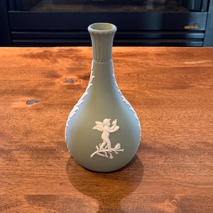 May include: A green ceramic vase with a white cherub design. The vase has a slender neck and a rounded body.