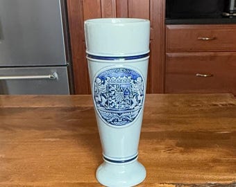 May include: A light blue ceramic beer stein with a flared base and a cylindrical body. The stein features a blue and white crest design with the word "BRYERN" in a circular banner. The stein is displayed on a wooden surface.