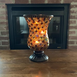 May include: A decorative mosaic glass candle holder with a flared shape. The candle holder is made of small, square tiles in shades of orange, gold, and brown. It has a dark brown base and rim, and is lit from within, creating a warm glow.