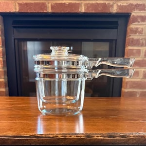 May include: A clear glass double boiler with a lid and two handles. The glass pot sits on a wooden surface in front of a brick fireplace.