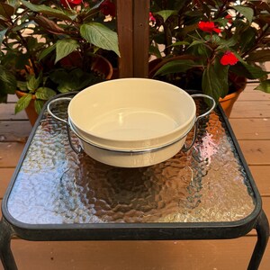 May include: A cream-colored, round serving bowl with a metal handle and rim. The bowl sits on a glass-topped table with a black metal frame. Potted plants with red flowers are in the background.