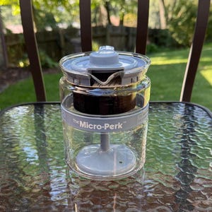 May include: A clear glass coffee percolator labeled The Micro-Perk with a light gray plastic lid, a central stem, and a dark brown internal basket. The cylindrical vessel features a pouring spout and a gray band around the upper rim.