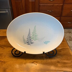 May include: An oval serving platter with a speckled, light blue and white surface. The platter features a fern design in shades of blue and green. The platter is displayed on a black metal stand.
