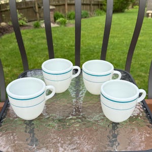 May include: Four white ceramic coffee cups with a teal blue stripe around the rim and a matching stripe around the middle. The cups are arranged on a glass table, with a green lawn in the background.