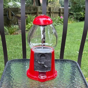 May include: A red gumball machine featuring a clear glass globe, a bright red lid, and a metallic gray coin mechanism with a rotating handle. The base is a vibrant red, and a small circular opening at the bottom allows for dispensing items.
